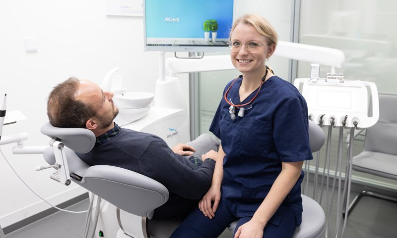 Oralchirurgin sitzt neben Patient und lächelt in die Kamera